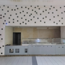 an empty stall in a mall food court. the facade is barren, just the tile pattern and font of the counter. there are open holes in drywall that show through to wiring that is no longer in use. the area behind the stall is empty, and unlit, and decaying. it's creepy.