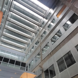 The interior of a tall, brutalist structure. The ceiling is several floors up, and many windows from other rooms overlook the camera, embedded in concrete walls. The ceiling itself has an odd shape to it, alternating glass and metal slats. At the edges of the image, strange lights just peek into frame.
