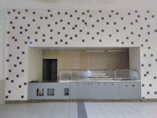 an empty stall in a mall food court. the facade is barren, just the tile pattern and font of the counter. there are open holes in drywall that show through to wiring that is no longer in use. the area behind the stall is empty, and unlit, and decaying. it's creepy.