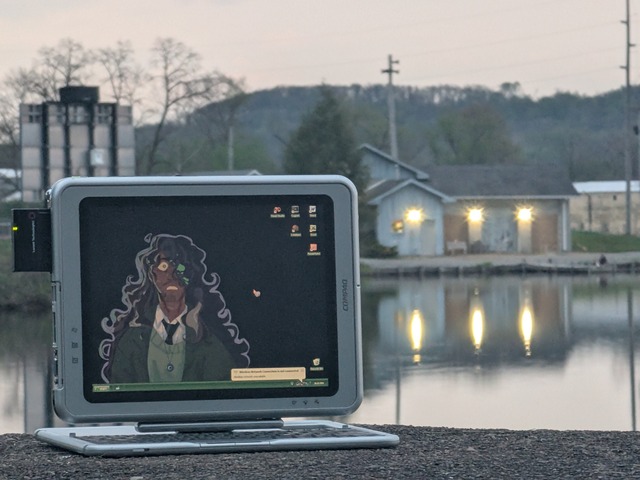 A compaq TC1000, by the water, at sunset. The wallpaper is Jonathan Sims, partially transformed into a monster. In the distance, there's a boathouse.
