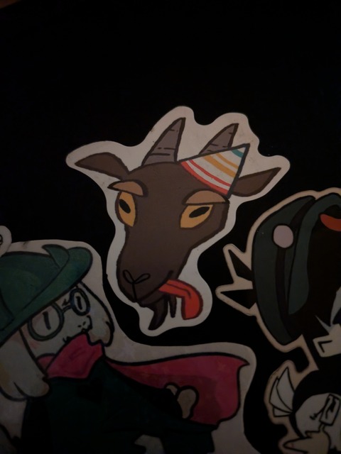A sticker of a goat head, in a party hat, sticking its tongue out. Under it is a ralsei sticker.