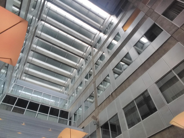 The interior of a tall, brutalist structure. The ceiling is several floors up, and many windows from other rooms overlook the camera, embedded in concrete walls. The ceiling itself has an odd shape to it, alternating glass and metal slats. At the edges of the image, strange lights just peek into frame.